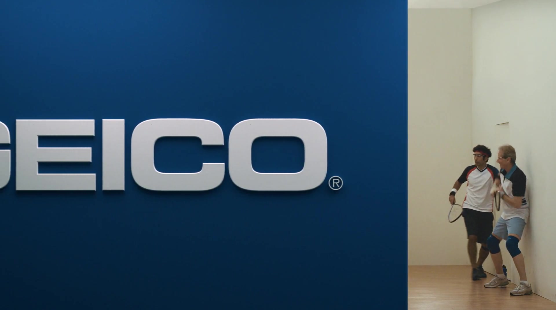 Two racquetball players squeeze against the court wall as a blue GEICO wall closes in.