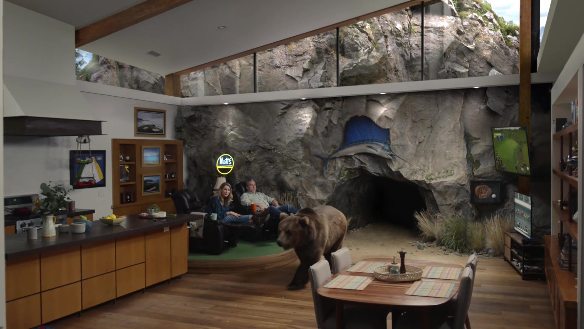 A bear walks from a rocky cave into an open kitchen while a couple sits in recliners.