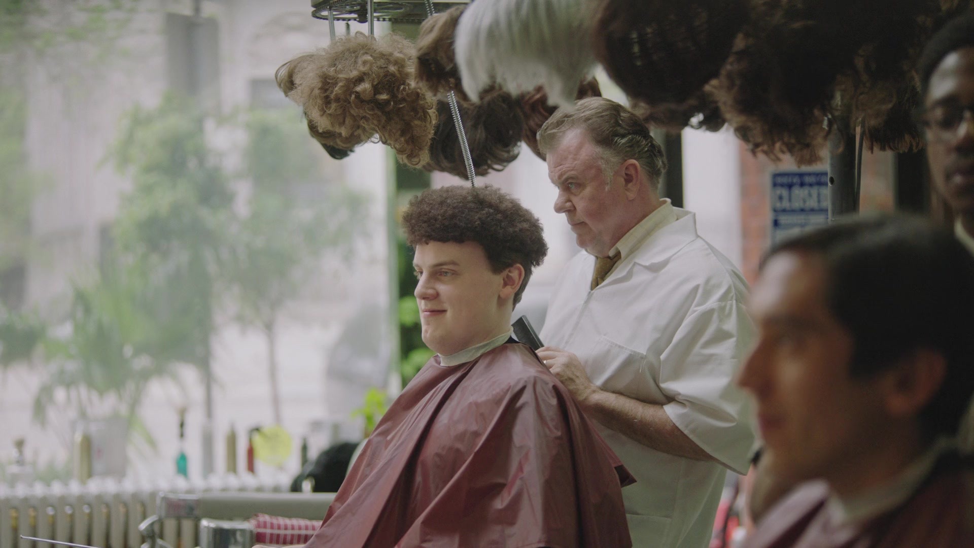 Customer tries another hairstyle beneath suspended wigs while barber watches.