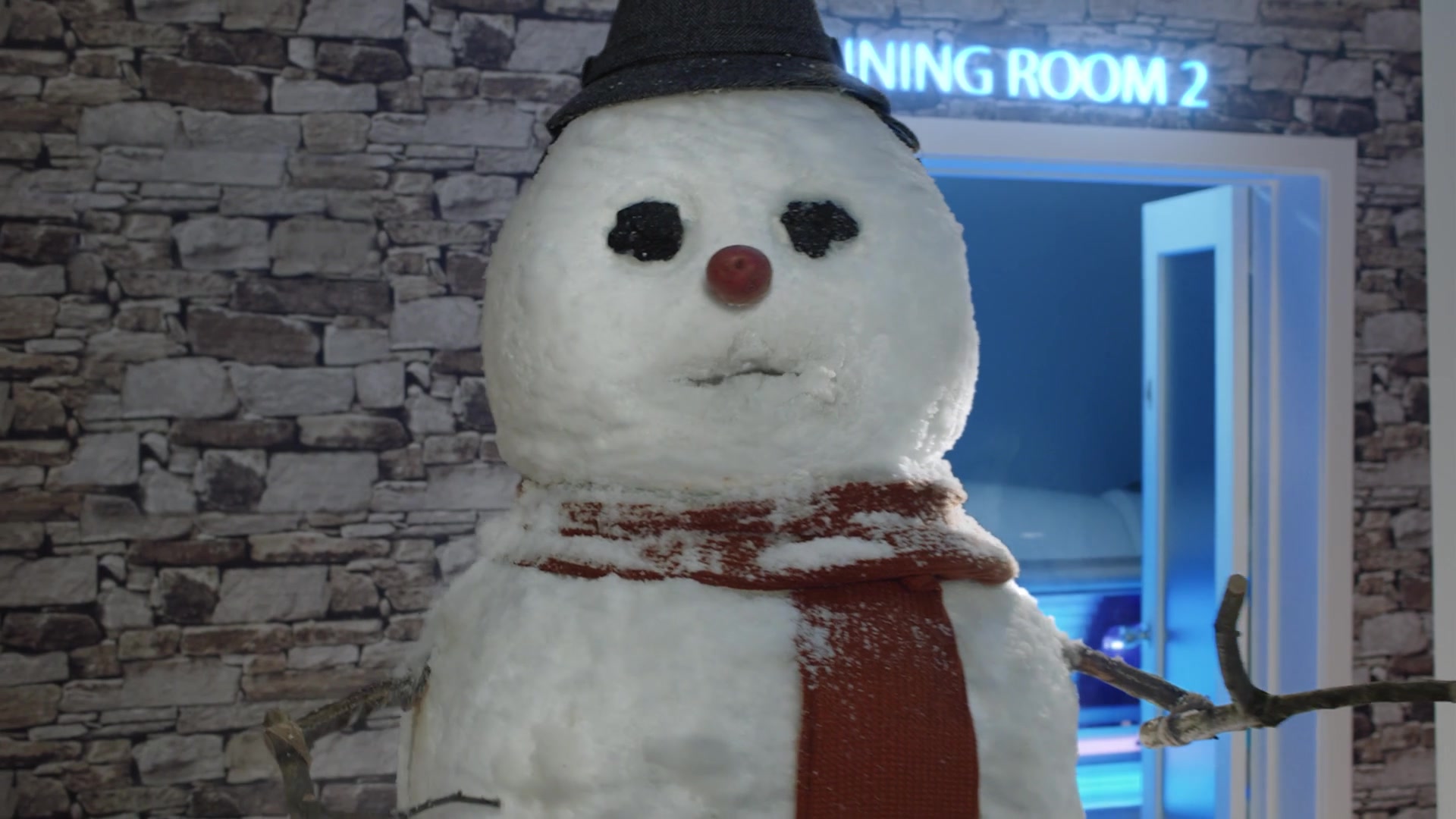 A snowman wearing a black hat and red scarf stands beside a glowing tanning room.