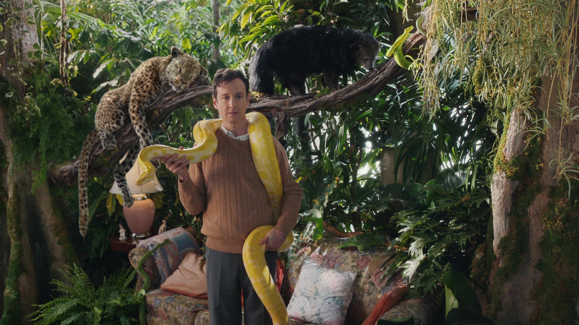 A man holds a yellow snake in a living room filled with tropical plants and wild animals.