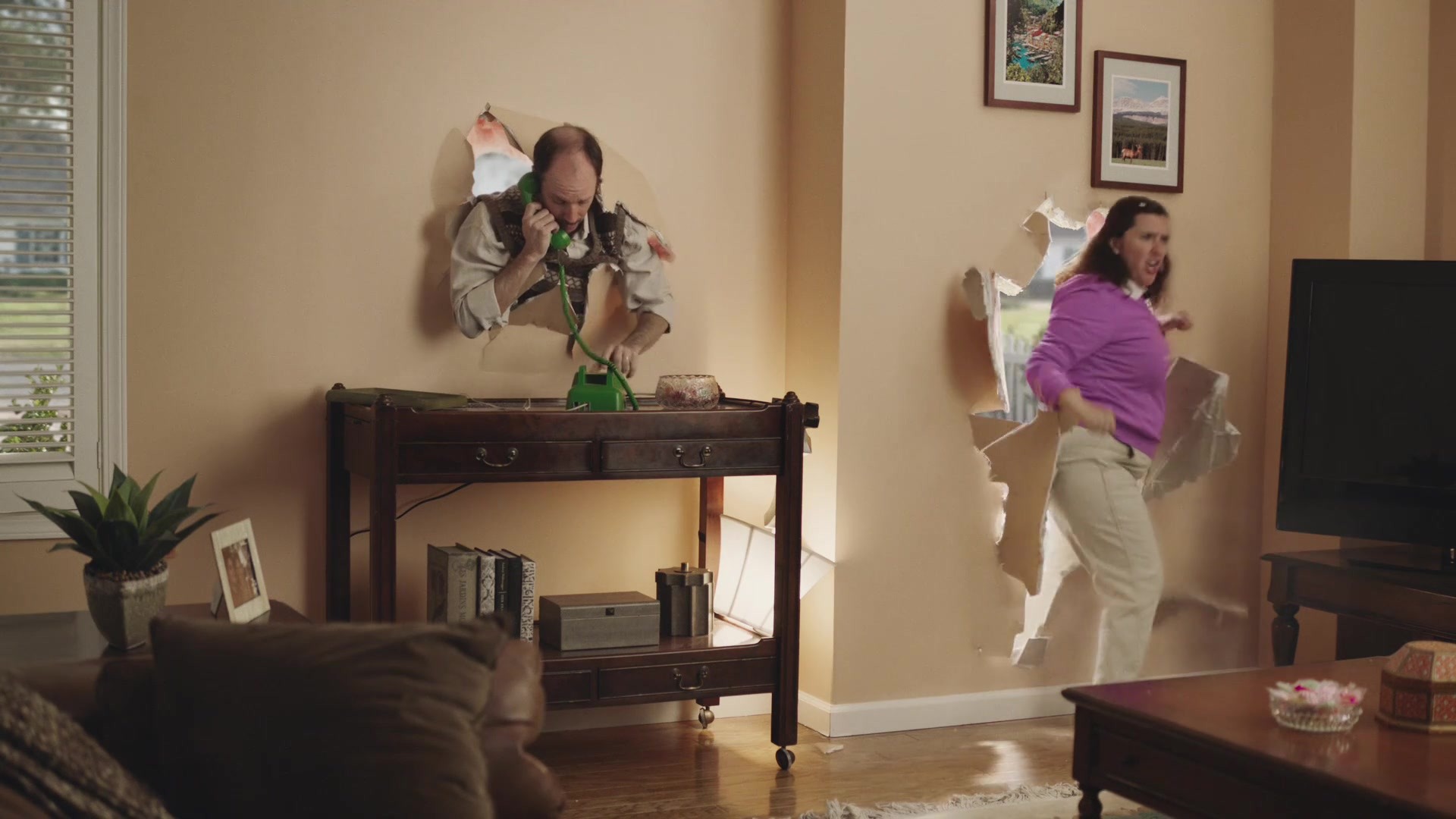 Wife bursts through living-room wall beside husband taking a phone call through his own hole.