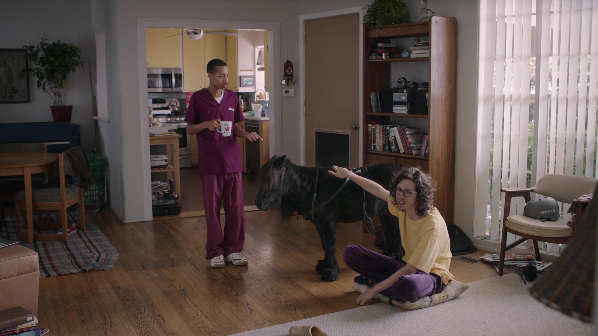 A man in scrubs watches his roommate pet a miniature horse in their living room.