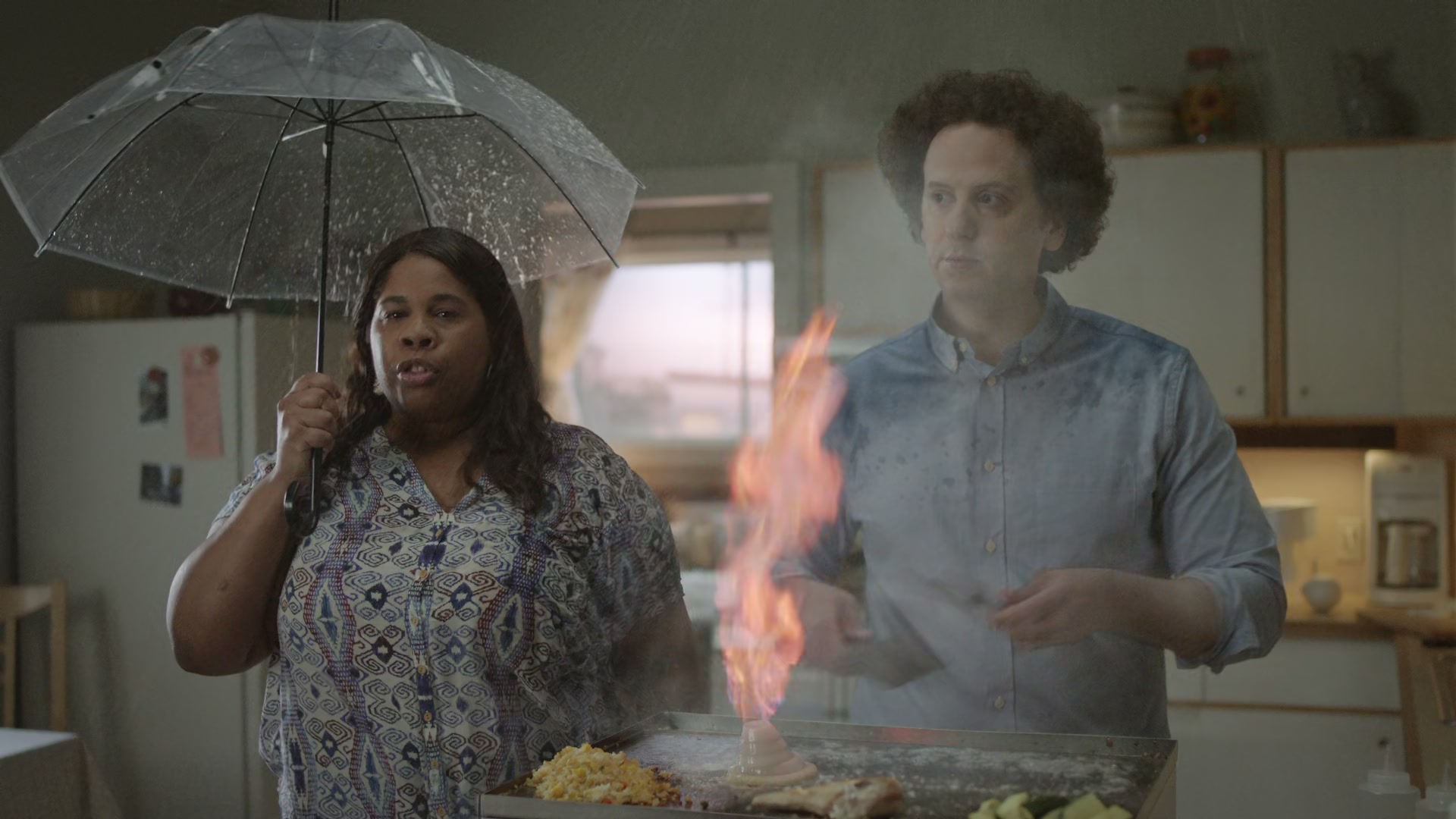 A woman holds an umbrella beside a man grilling a flaming onion tower as sprinklers soak the kitchen.
