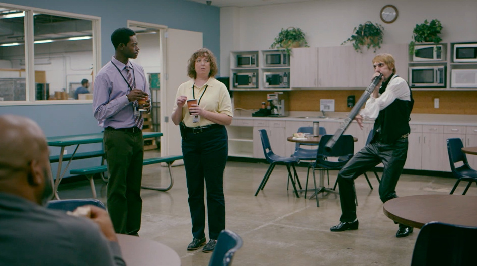A didgeridoo player performs beside two coworkers holding cups in an office break room.