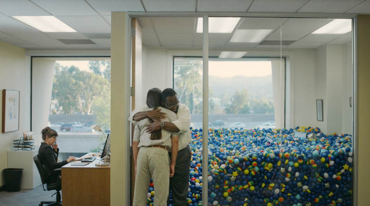 Two coworkers hug beside a glass-walled office filled with colorful stress balls.